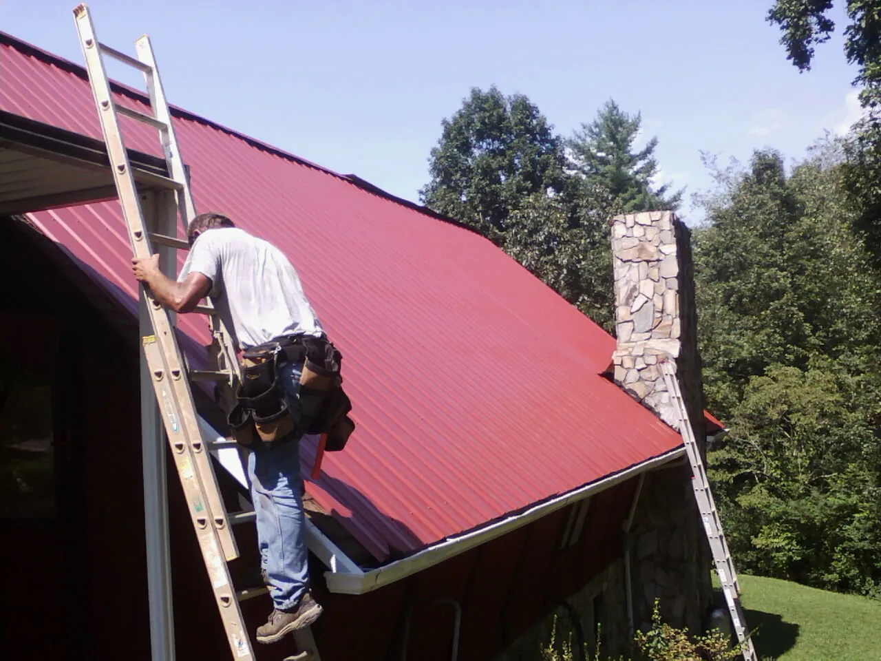 Roofer on a ladder beside a red metal roof and stone chimney — Soffit & Fascia Repair by Roofing Brothers in Vickery