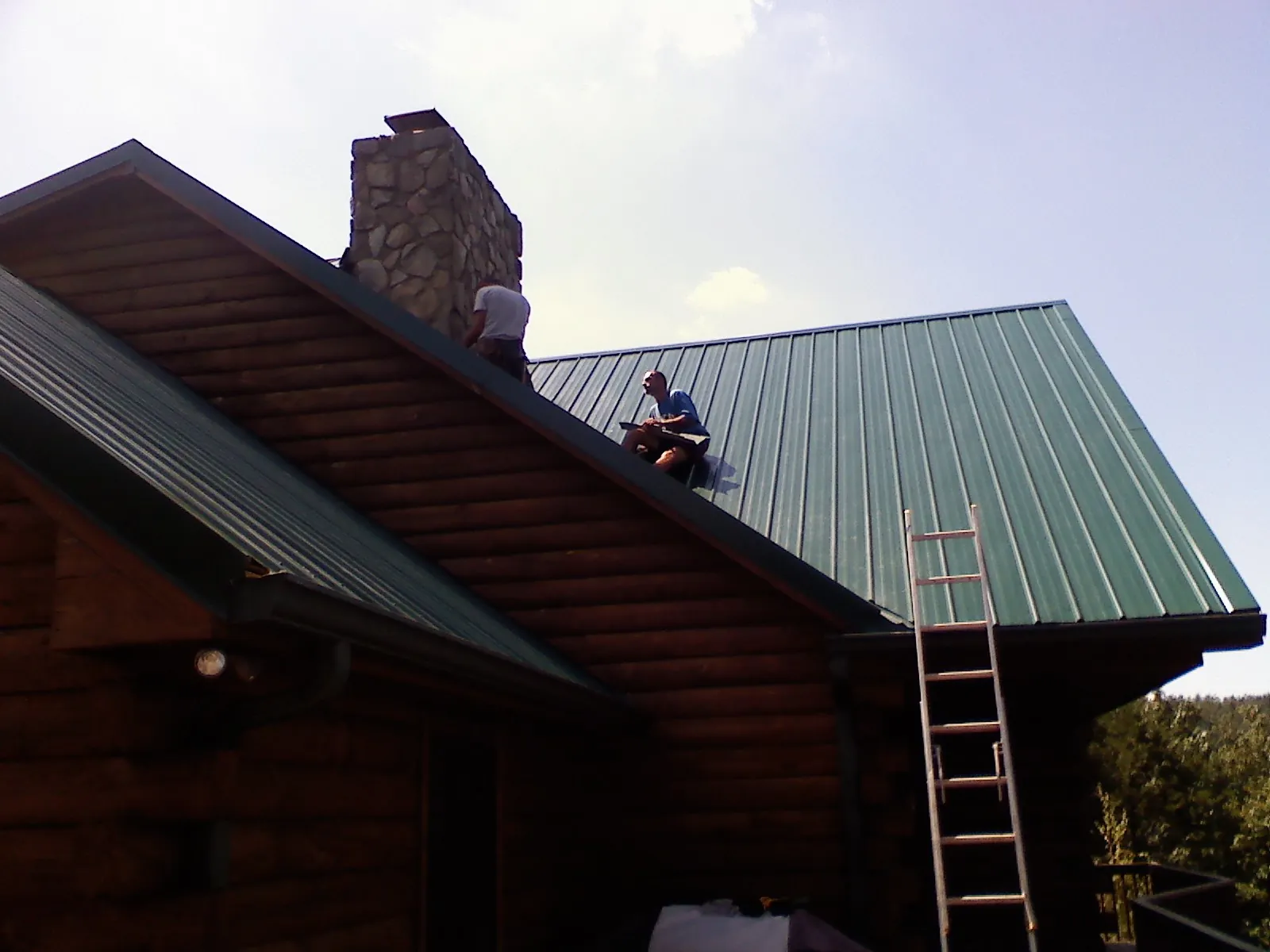 Roofers installing green metal panels beside a stone chimney on a log home in Vickery